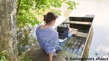 A woman sits by a lake, working on her laptop