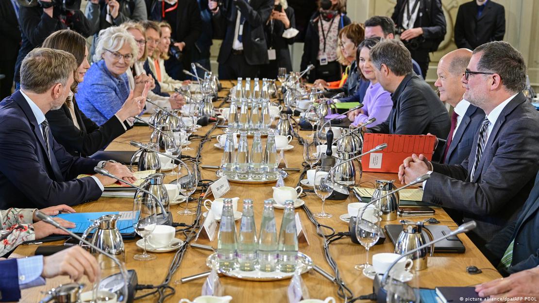 Líderes do governo alemão estão sentados a uma extensa mesa, com microfones individuais, pastas e garrafas d'água ao centro.