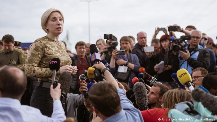 A woman wearing a camouflage top speaks to a circle of reporters with microphones and cameras