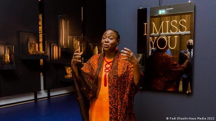 Peju Layiwola, a woman in an orange dress gestures as she speaks in front of a mirror with the words 
