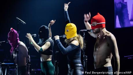 Four people wearing black suits and balaclavas singing into microphones.