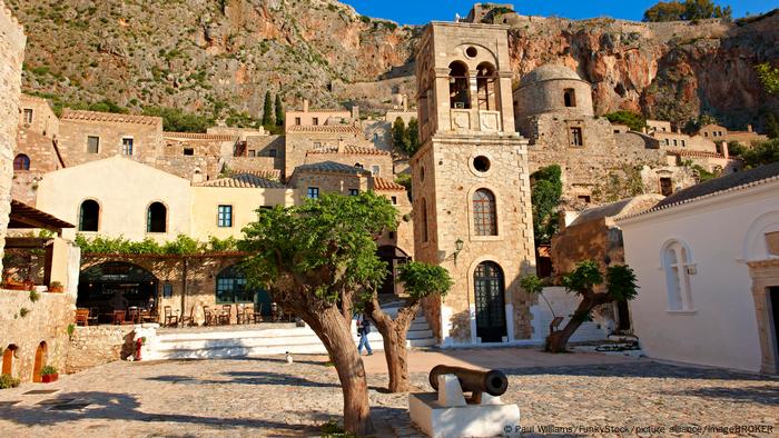 Buildings in the city of Monemvasia in front of a rocky cliff with stone houses and trees in a square.