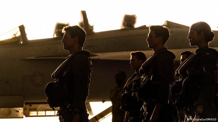 A group of uniformed pilots standing and in the background is a fighter jet