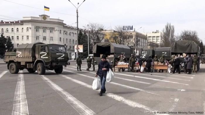 A Russian soldier carrying two bags of humanitarian aid; behind him are other soldiers and military vehicles marked with white 'Zs'