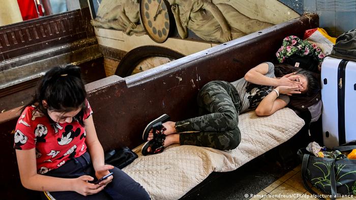 Zwei ukrainische Frauen in einer Notunterkunft für Flüchtlinge in Prag: eine liegt auf einer Bank und hält sich die Hände vors Gesicht, die andere sitzt neben ihr und schaut auf ihr Mobil-Telefon