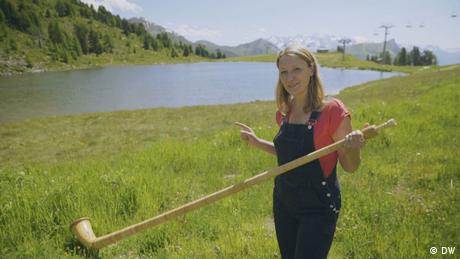 Eine Frau steht vor einem See in den Alpen und hält ein Alphorn in der Hand. (Bildrechte: DW)