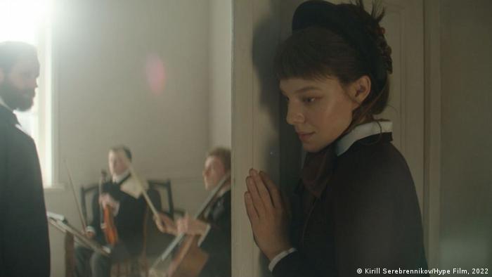 Scene from a film: a young white woman with a black dress and white collar and cuff is listening at a door; in the room behind is a man with a beard, a cellist and a violinist are sitting in front of him