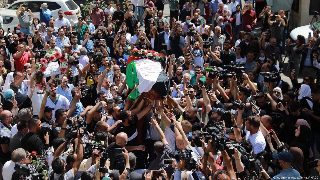 Homens carregam o caixão coberto com a bandeira palestina. Muitas pessoas no entorno, cinsluindo repórteres.