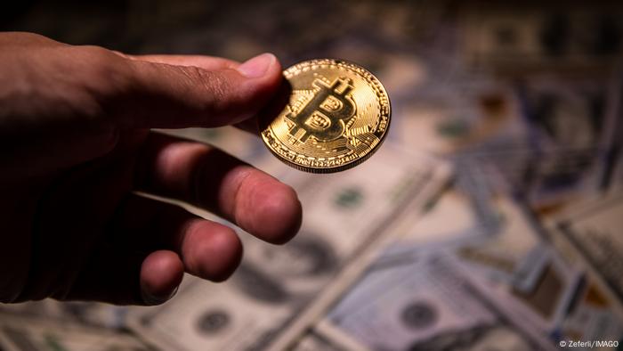 Eine Hand halt eine Bitcoin-Symbolmünze, im Hintergrund liegen Dollarscheine 