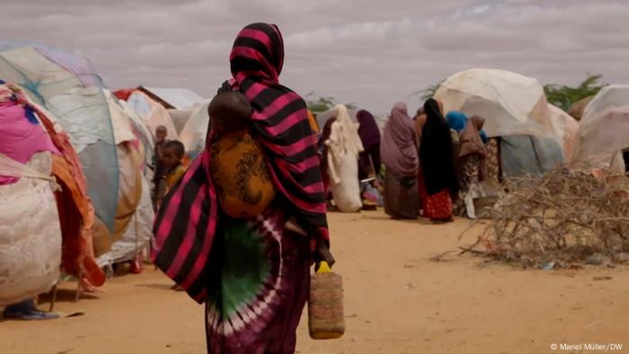 Woman in the foreground with a child on her back and several other women amid tents