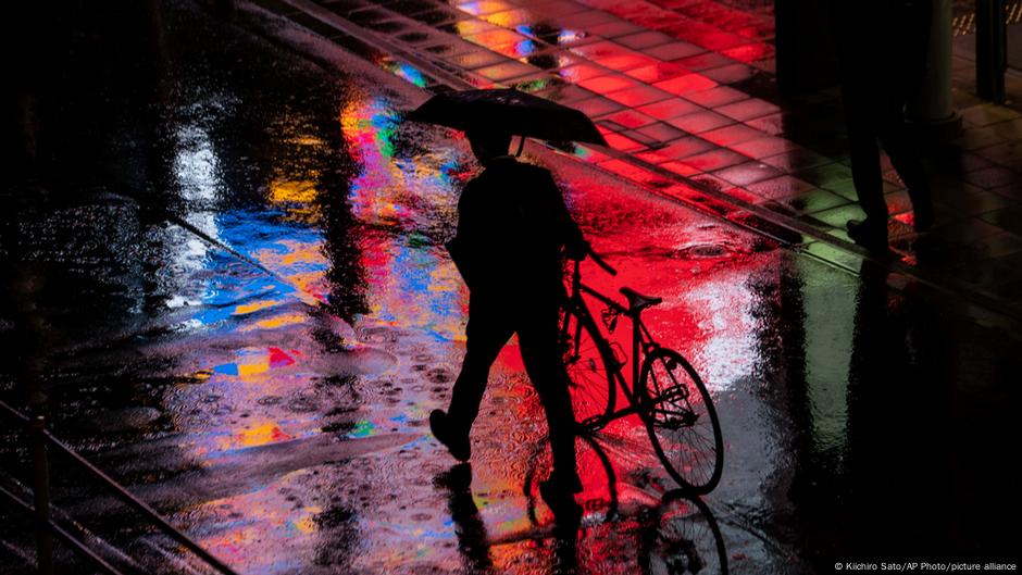 A man walks his bicycle in the rain, at night, as colorful lights reflect in the puddles