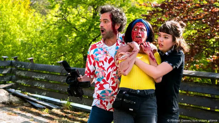 Eine Filmszene aus Coupez!: ein Mann im Hawaiihemd, eine Frau mit gelbem Pullover, blutverschmiertem Gesicht und weitgeöffneten Augen, die von einer weiteren jungen Frau gehalten wird