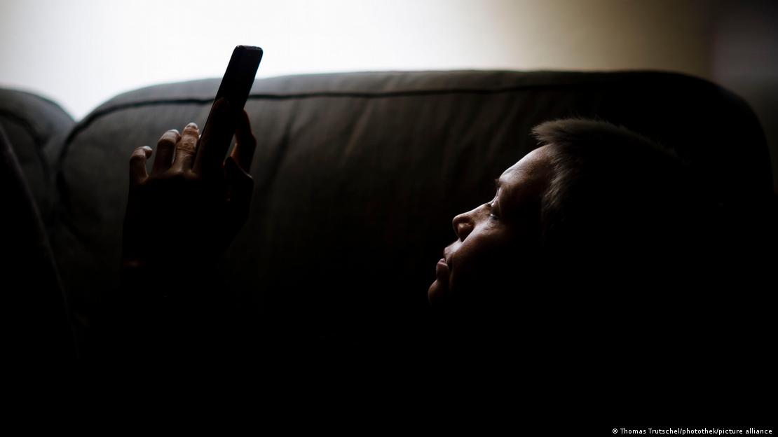 Pessoa deitada em sofá, em local escuro, observa celular. 