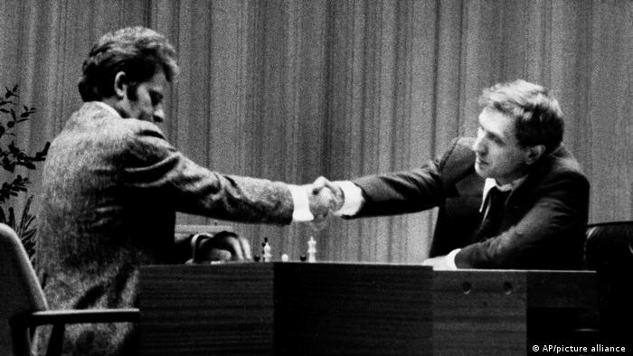 Schwarz-weiß-Aufnahme von Boris Spasski und Bobby Fischer, die sich am Schachtisch die Hände schütteln. 