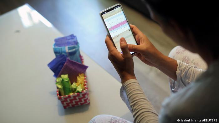 A woman in Spain, photographed from the back, sits while looking at a period calendar tracker on a smartphone. In front of her on a table is a small basket of tampons and other menstrual aids. 