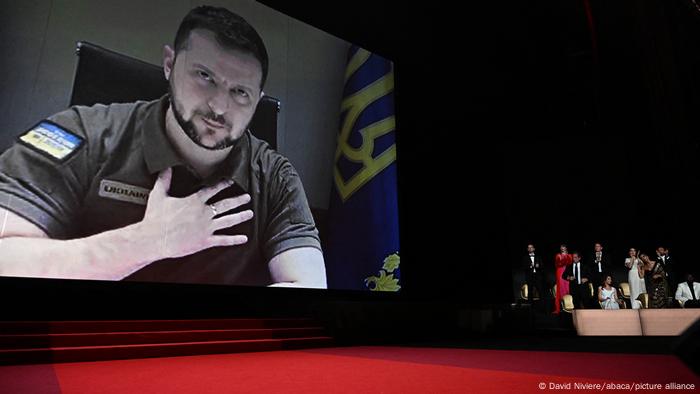 Ukrainian President Volodymyr Zelenskyy speaks at the Cannes film festival on a large screen; he is holding his hand over his chest and has a Ukrainian flag patch on his sleeve