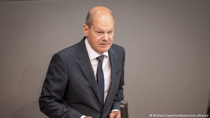 Olaf Scholz wears a grey suit and stands with slightly hunched posture