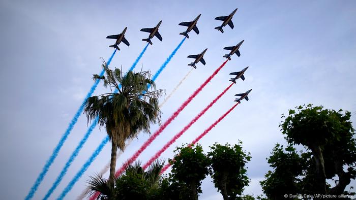 Mavi, beyaz ve kırmızı duman izleri olan jet avcı uçakları bir palmiye ağacının üzerinde uçuyor.