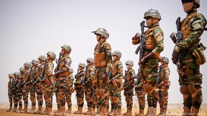 Several soldiers stand in a desert, wearing bandannas over their mouths