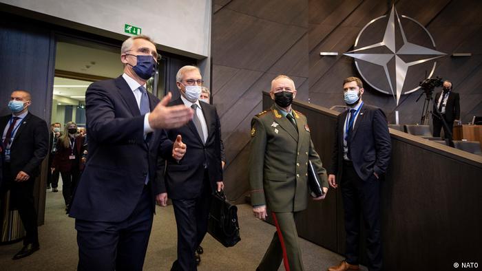 Mavi takım elbiseli ve biri yeşil Rus ordusu üniformalı iki adam, hepsi yüz maskeleri takmış, arkalarındaki duvarda NATO sembolü olan bir odaya giriyorlar.