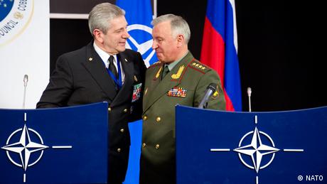 Two men in uniform pat each other on the back. One is wearing a navy-blue unform and standing in front of a NATO flag, one a green uniform in front of a Russian flag.