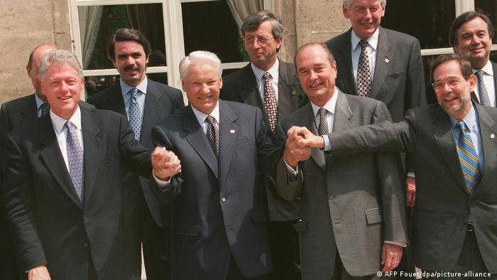Soldan: Bill Clinton, Boris Yeltsin, Jacques Chirac ve Javier Solana bir binanın önünde duruyor, el ele tutuşuyor ve sırıtıyor. Diğer NATO liderleri de arkalarında duruyor. Dokuz liderin hepsi erkek ve hepsi gri takım elbise giyiyor.