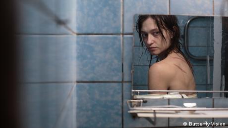 A still from 'Butterfly Vision': a mirror reflection of a woman looking at wounds on her back, in a blue-tiled bathroom.