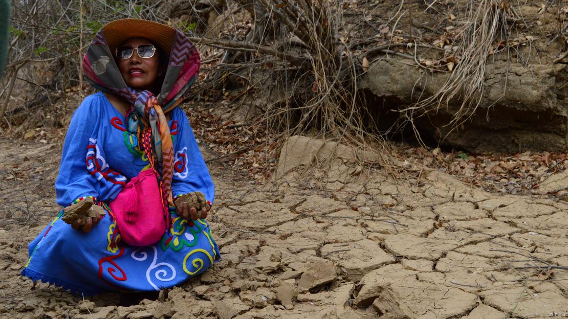 Mulher sentada sobre leito de rio seco, segurando pedaços da terra seca na mão