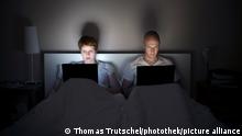 A young couple sit in bed in the dark and work on their laptops