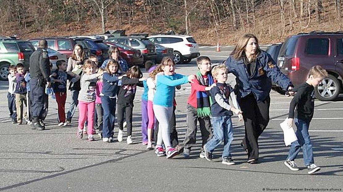 A imagem mostra uma fila de crianças assustadas, junto a professoras e um policial, em um estacionamento próximo à escola onde um atentado com arma de fogo havia ocorrido, em 14 de dezembro de 2012, em Newtown, nos Estados Unidos.