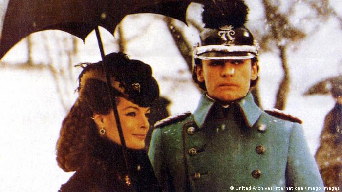 Romy Schneider und Helmut Berger im Film Ludwig II. Die beiden Darsteller stehen nebeneinander, er guckt ernst, sie lächelt. 