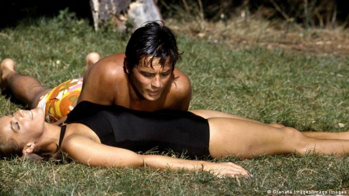 Romy Schneider in a black bathing suit lies on the grass with actor Alain Delon 