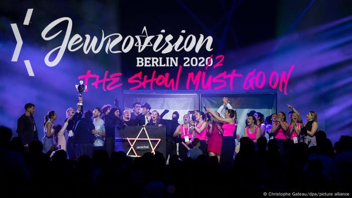 Die Gewinner des Jewrovision Song Contests 2022 stehen auf der Bühne, über ihnen der Schriftzug Jewrovision. Berlin 2022 - The show must go on. Im Vordergrund die Silhouetten der Zuschauer.