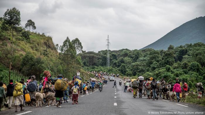 A long line of people carrying goods and pulling animals walk along a road