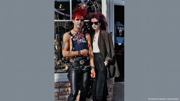 Ein Punk-Pärchen mit gefärbten Haaren vor einem Geschäft, er trägt Nietenklamotten und Muscle Shirt, sie Sonnenbrille, beide haben dei Gesicher weiß geschminkt.
