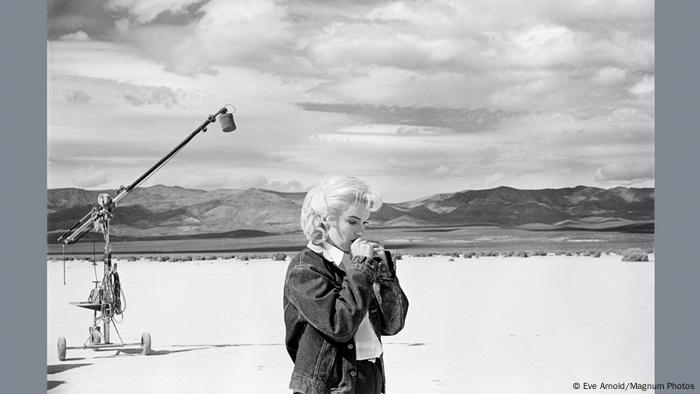 Marilyn Monroe steht, sichtlich konzentriert, auf einem Filmset vor einer weiten Wüstenlandschaft mit einem Sound-Kran.