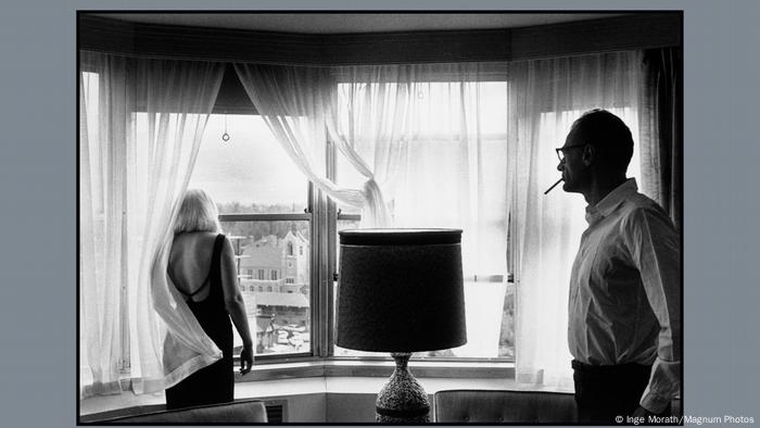 Marilyn Monroe blickt aus einem Fenster, ihr Rücken ist zu sehen, im Vordergrund im Profil Arthur Miller mit Zigarette im Mund. Er blick zu ihr hinüber.