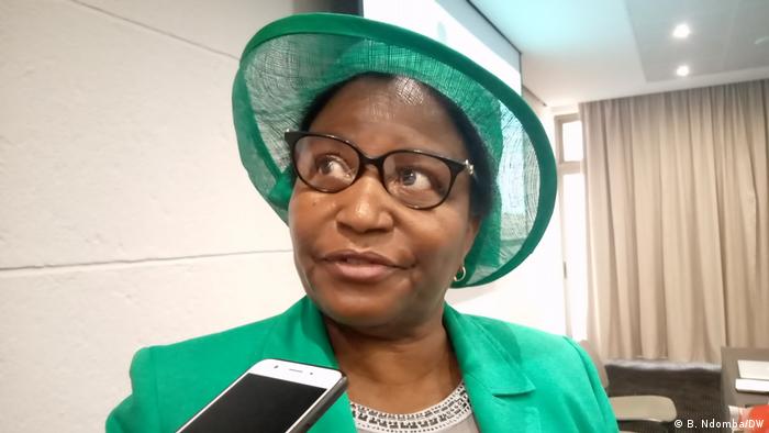 Woman in a green hat and jacket in interview. A cellphone is held at the ready to record her answers