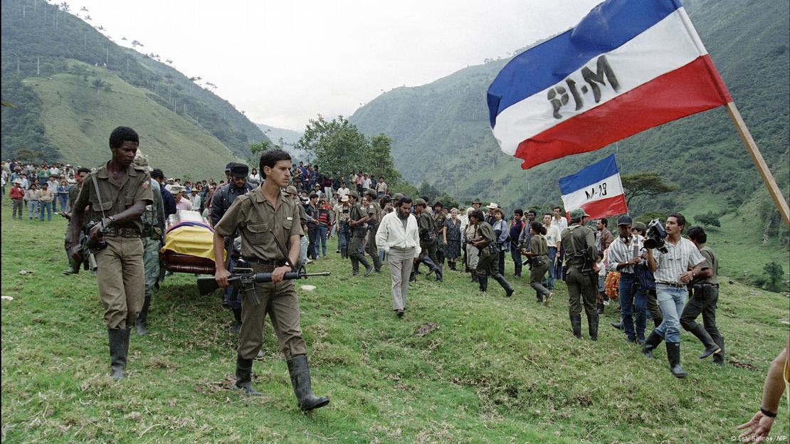 Homens fardados e armados carregando um caixão em uma área montanhosa 