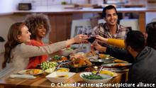 Side view of a group of young adult multi-ethnic male and female friends sitting at a table at home set for Thanksgiving dinner making a toast with glasses of red wine || Modellfreigabe vorhanden