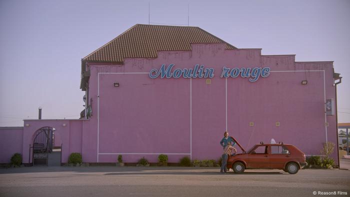 In einer Szene des Films My Thoughts Are Silent stehen zwei Männer am Motor eines roten Autos, das vor einem pinkfarbenen Haus steht.