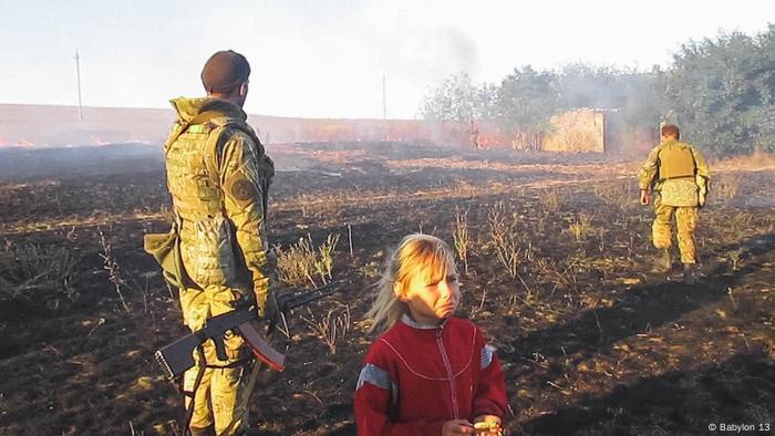 In einer Szene des Films War Note stehen zwei Soldaten mit einem traurigen Kind vor einem brennenden Feld.