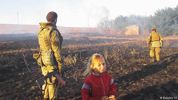 In einer Szene des Films War Note stehen zwei Soldaten mit einem traurigen Kind vor einem brennenden Feld.