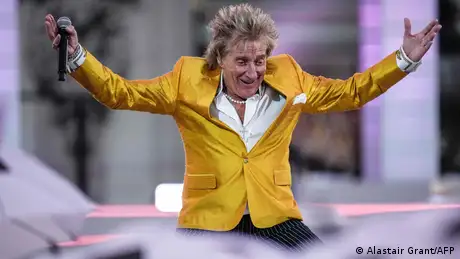 Rod Stewart holding a microphone, raises his arms as his smiles, wearing a yellow suit.