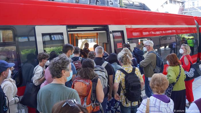 Menschen stehen gedrängt an, um in einen Zug einzusteigen. Sie sind sommerlich gekleidet, einige tragen einen Mund-Nasen-Schutz. Der Zug ist rot, die Türen sind geöffnet. Menschen stehen gedrängt an, um in einen Zug einzusteigen. Sie sind sommerlich gekleidet, einige tragen einen Mund-Nasen-Schutz. Der Zug ist rot, die Türen sind geöffnet.