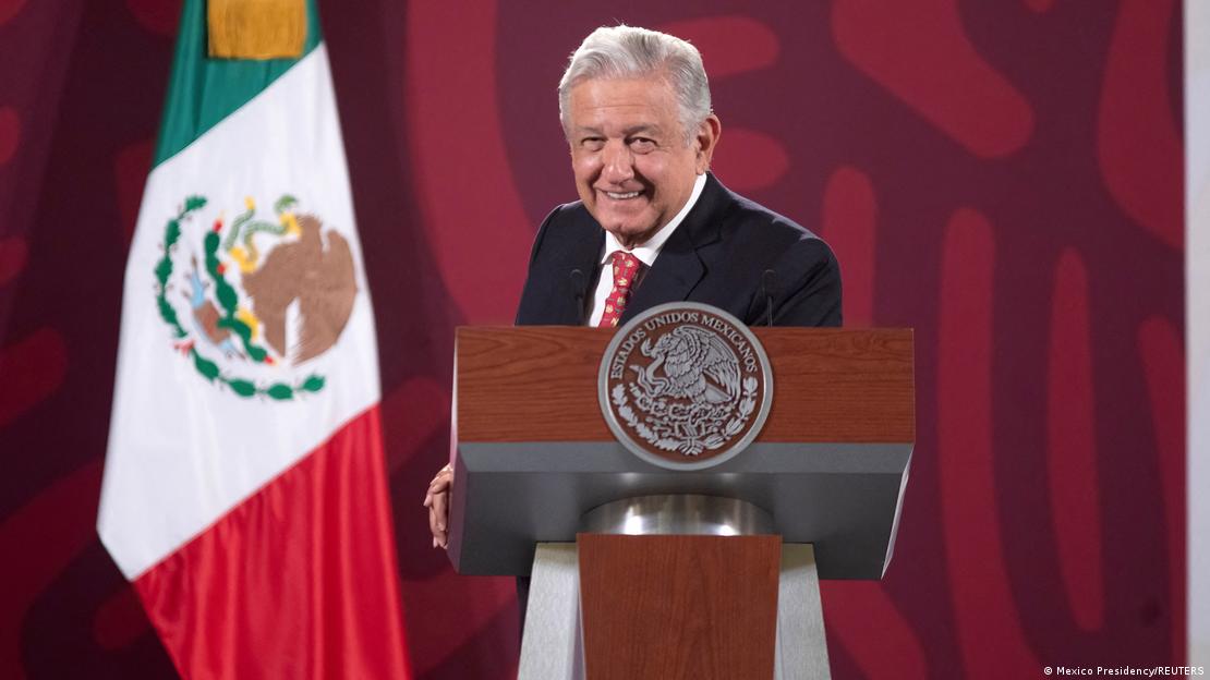A foto mostra o presidente mexicano, Andres Manuel Lopez Obrador. Ele veste terno escuro, camisa branca e gravata vermelha, e está atrás de um púlpito oficial do governo mexicano. Ao fundo, à esquerda, está a bandeira do México.