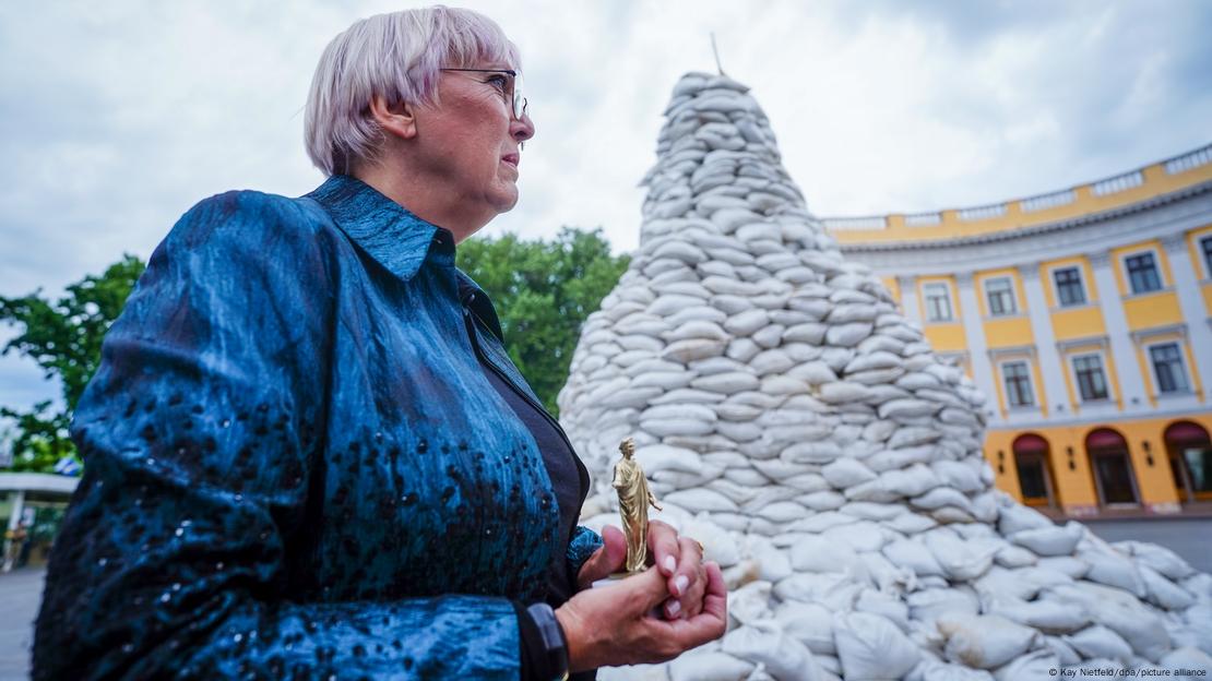 Kulturstaatsministerin Roth steht neben einem mit Sandsäcken geschützten Monument in Odessa und hält eine goldene Figur in den Händen. 