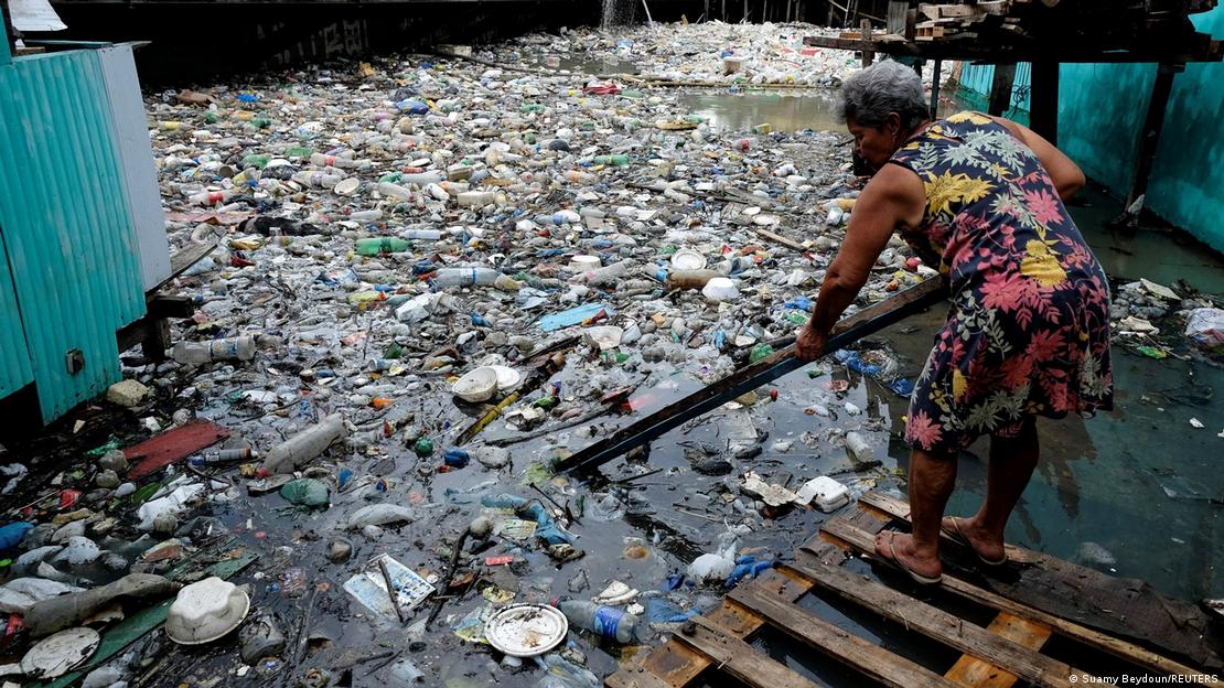 Mulher procura alumínio em rio em Manaus, usando máscara e de pé num pallet de madeira, cavoucando a água com um pedaço de madeira