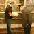 Ein älterer und ein jüngerer Mann werfen in einer Bibliothek einen Blick in ein Buch, das der jüngere in den Händen hält.