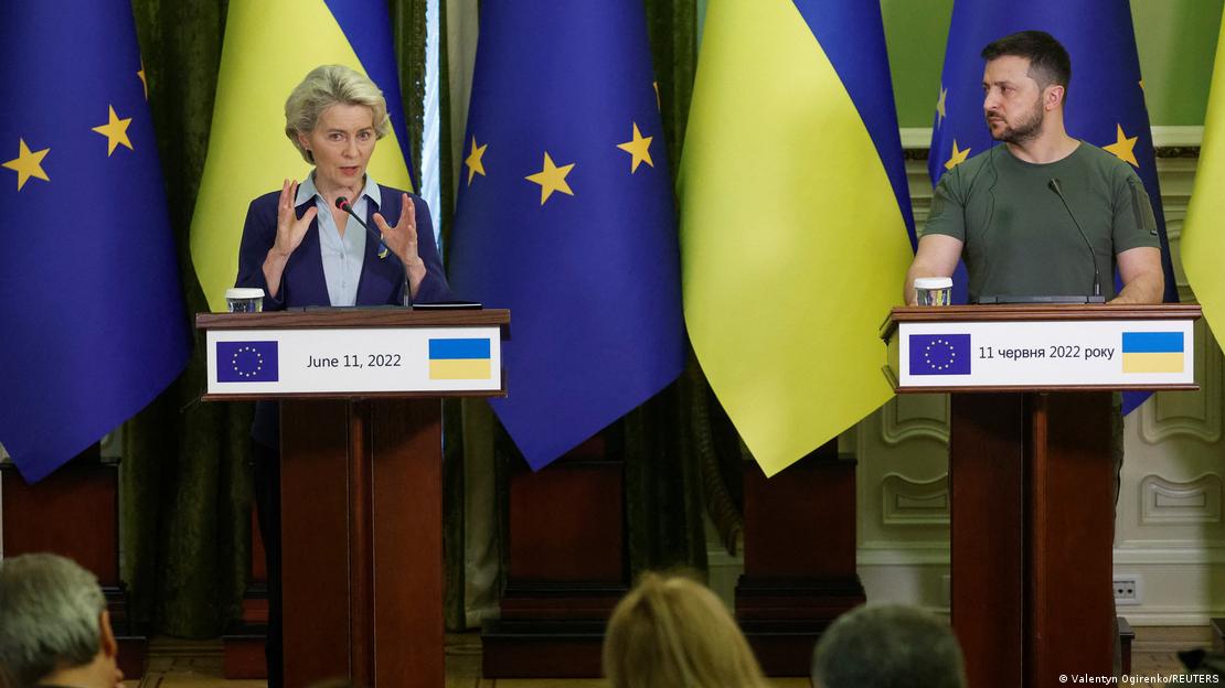 A imagem mostra a presidente da Comissão Europeia, Ursula von der Leyen, à esquerda, e o presidente da Ucrânia, Volodimir Zelenski, à direita. Eles estão em um palco, atrás de um púlpito e à frente de bandeiras da União Europeia e da Ucrânia, e dão explicações para a imprensa.